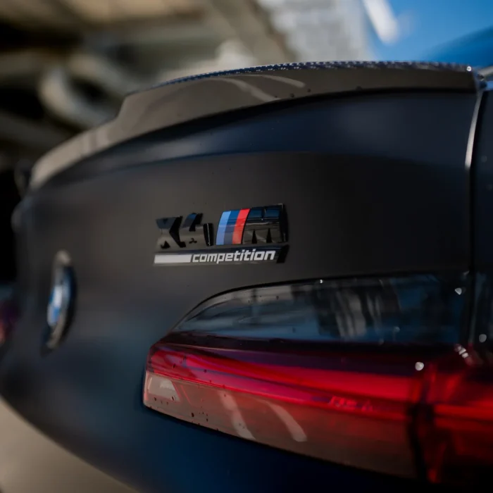 Close-up of BMW X4M Competition badge and carbon lip spoiler, highlighting M branding and carbon weave